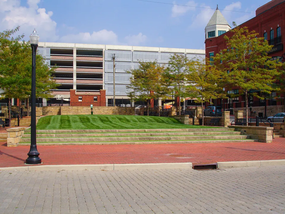 Tiered lawn panel and brick pedestrian paving beside a parking structure and commercial buildings.